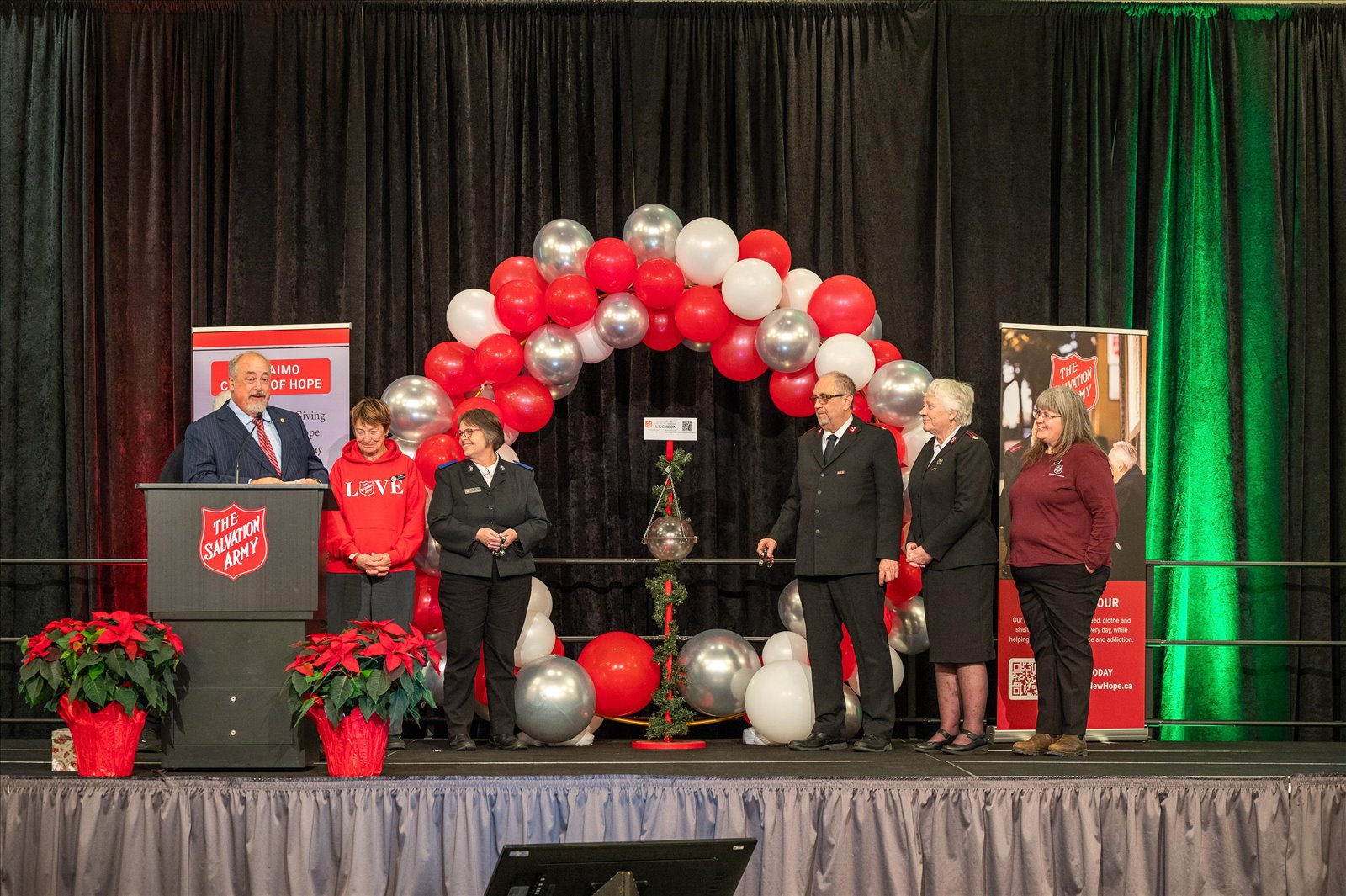 The Salvation Army representatives standing on a stage during the Hope in the Harbour event, with a speaker at a podium and red, white, and silver balloon decorations.